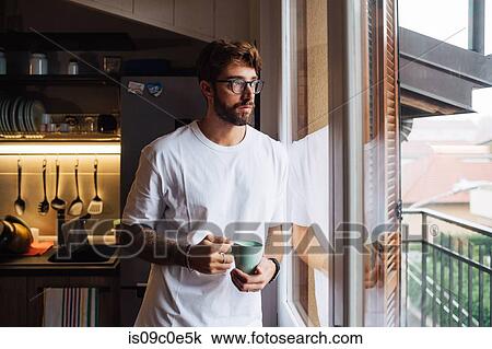 Stock Image - Mid adult man with coffee cup gazing out through apartment window. Fotosearch