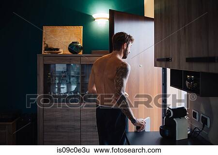 Stock Photograph - Mid adult man with tattooed arm leaving kitchen, rear view. Fotosearch