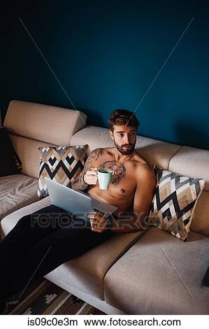 Picture - Mid adult man with tattooed chest on sofa with laptop and coffee. Fotosearch