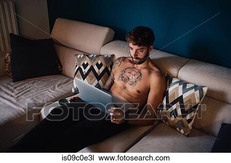 Mid adult man with tattooed chest on sofa looking at laptop View Large Photo Image Stock Image - Mid adult man with tattooed chest on sofa looking at laptop. Fotosearch