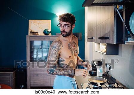 Stock Image - Mid adult man with tattooed chest preparing breakfast while looking over shoulder. Fotosearch