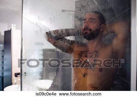 Stock Image - Mid adult man with tattooed chest taking a shower with eyes closed. Fotosearch