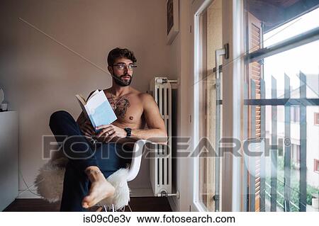 Stock Photo - Mid adult man with tattooed chest sitting on chair looking out through window. Fotosearch