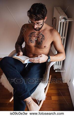 Stock Photo - Mid adult man with tattooed chest sitting on chair reading book. Fotosearch