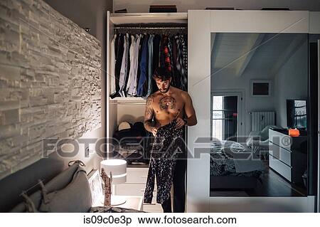 Stock Photography - Mid adult man with tattooed chest getting shirt from wardrobe. Fotosearch