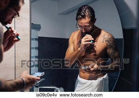 Stock Photo - Mid adult man with tattoos brushing teeth and looking at smartphone at bathroom mirror, mirror image. Fotosearch