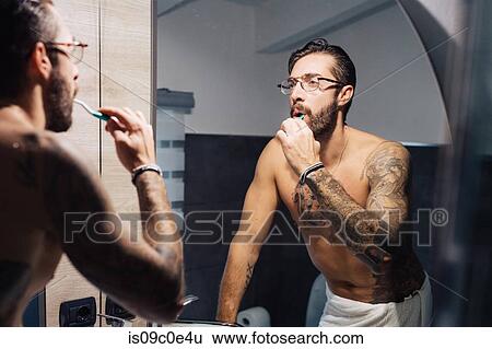 Stock Photograph - Mid adult man with tattoos brushing teeth at bathroom mirror. Fotosearch
