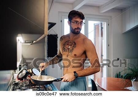 Stock Photo - Mid adult man with tattoos cooking breakfast in frying pan. Fotosearch