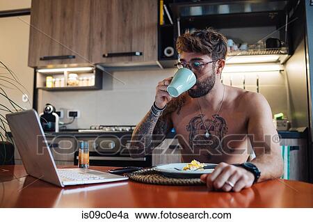 Stock Image - Mid adult man with tattoos drinking coffee while looking at laptop. Fotosearch