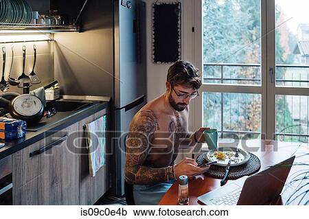 Stock Image - Mid adult man with tattoos drinking coffee while looking at smartphone. Fotosearch
