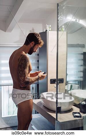 Stock Photo - Mid adult man with tattoos looking at smartphone at bathroom mirror. Fotosearch