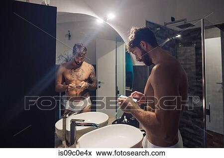 Stock Photo - Mid adult man with tattoos using smartphone touchscreen at bathroom mirror, mirror image. Fotosearch