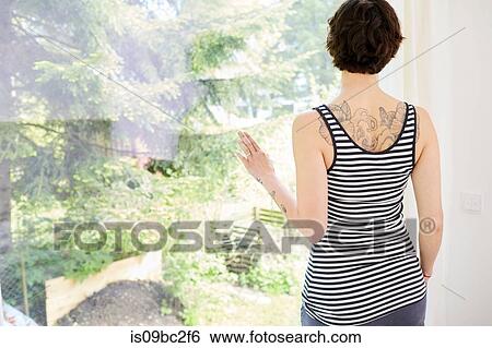 Stock Photograph - Mid adult woman, at home, looking out of window, rear view. Fotosearch