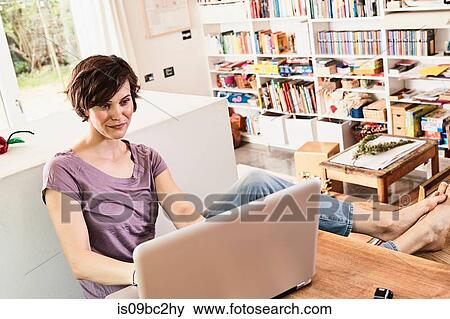 Stock Photo - Mid adult woman at home, sitting it table, using laptop. Fotosearch