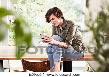 Mid adult woman at home, sitting on table, using smartphone View Large Photo Image Picture - Mid adult woman at home, sitting on table, using smartphone. Fotosearch