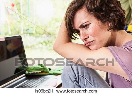 Stock Image - Mid adult woman at home, using laptop, worried expression. Fotosearch