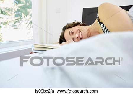 Stock Image - Mid adult woman, relaxing on bed, holding book, smiling. Fotosearch