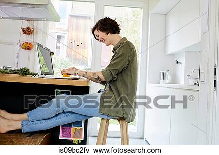 Mid adult woman sitting in kitchen, using laptop, laughing View Large Photo Image Stock Photo - Mid adult woman sitting in kitchen, using laptop, laughing. Fotosearch