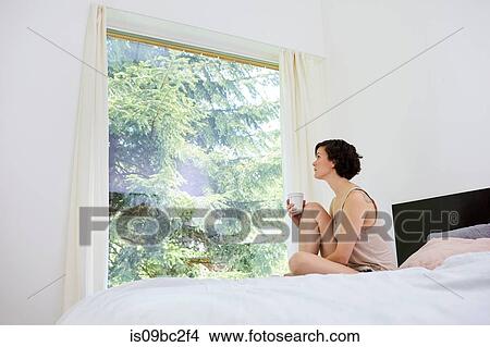 Mid adult woman, sitting on bed, holding coffee cup, looking out of window View Large Photo Image Picture - Mid adult woman, sitting on bed, holding coffee cup, looking out of window. Fotosearch