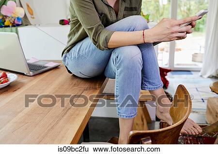 Stock Photograph - Mid adult woman sitting on kitchen table, using smartphone, low section. Fotosearch