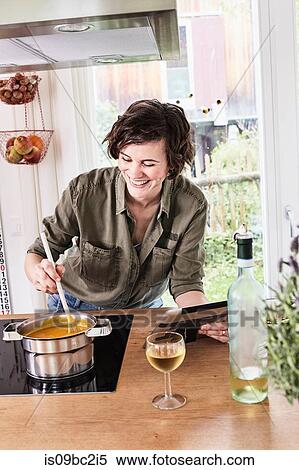Stock Photography - Mid adult woman stirring pot on stove in kitchen, holding digital tablet, laughing. Fotosearch