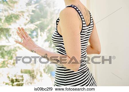 Stock Photo - Mid adult woman, with tattoos on arm, looking out of window, mid section. Fotosearch