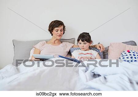 Mother and daughter relaxing in bed, daughter reading book, mother holding digital tablet View Large Photo Image Stock Photo - Mother and daughter relaxing in bed, daughter reading book, mother holding digital tablet. Fotosearch