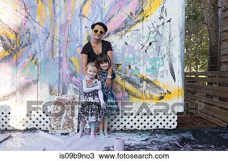 Stock Image - Mother and daughters covered in paint by painted wall. Fotosearch