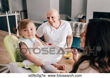 Mother and father feeding baby daughter in child seat on kitchen table View Large Photo Image Stock Photo - Mother and father feeding baby daughter in child seat on kitchen table. Fotosearch