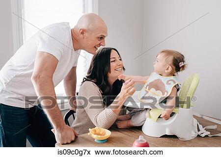 Mother and father feeding baby daughter in child seat on kitchen table View Large Photo Image Stock Photo - Mother and father feeding baby daughter in child seat on kitchen table. Fotosearch