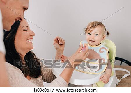 Stock Photo - Mother and father feeding baby daughter in child seat, portrait. Fotosearch