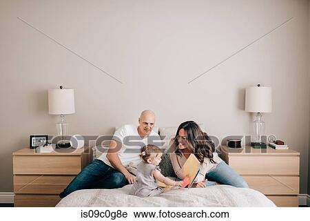 Stock Image - Mother and father on bed with baby daughter looking at story book. Fotosearch