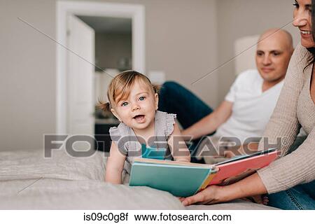 Stock Image - Mother and father on bed with baby daughter with story book, portrait. Fotosearch