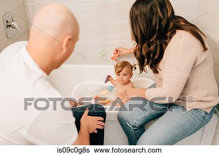 Stock Photograph - Mother and father playing with baby daughter in bath, over shoulder view. Fotosearch