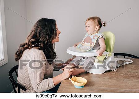 Mother feeding baby daughter in child seat on kitchen table View Large Photo Image Stock Photo - Mother feeding baby daughter in child seat on kitchen table. Fotosearch