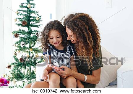 Stock Image - Mother giving daughter present beside Christmas tree at home. Fotosearch