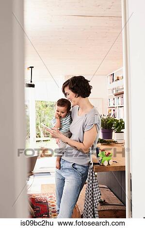 Mother holding baby daughter in kitchen, looking at smartphone View Large Photo Image Stock Photograph - Mother holding baby daughter in kitchen, looking at smartphone. Fotosearch