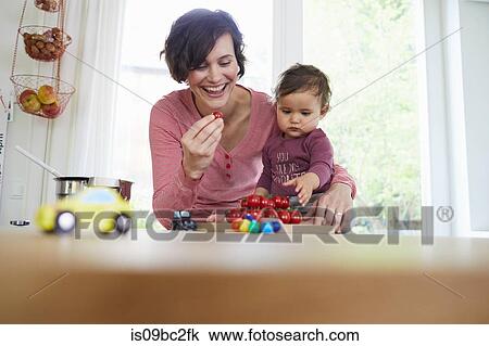 Stock Image - Mother holding baby girl at kitchen counter, pile of tomatoes in front of them. Fotosearch