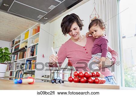 Stock Image - Mother holding baby girl in kitchen, whilst stirring pot on stove. Fotosearch