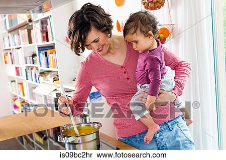 Stock Photography - Mother holding baby girl in kitchen, whilst stirring pot on stove. Fotosearch