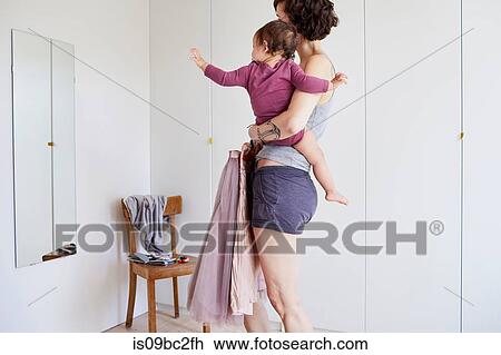 Stock Image - Mother holding baby girl, whilst looking in mirror, deciding what to wear. Fotosearch