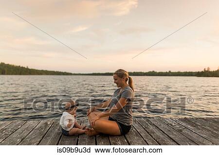 Stock Image - Mother sitting cross legged on lake pier with baby daughter. Fotosearch