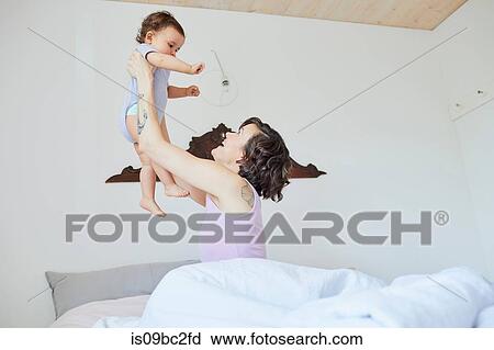 Stock Image - Mother sitting in bed, holding baby girl in air, smiling. Fotosearch