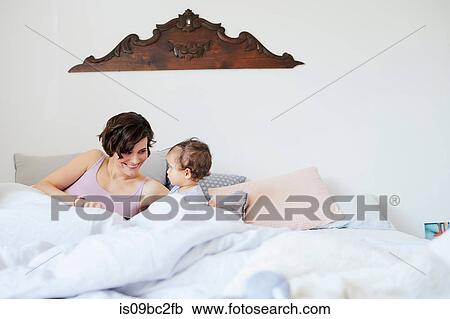 Stock Image - Mother sitting in bed with baby girl, smiling. Fotosearch