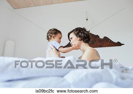 Stock Image - Mother sitting in bed with baby girl, face to face, pensive expression. Fotosearch
