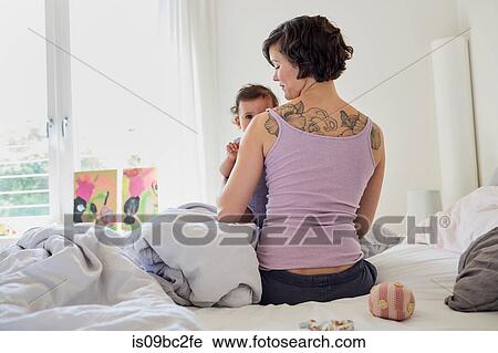 Stock Image - Mother sitting on bed, holding baby girl, rear view. Fotosearch