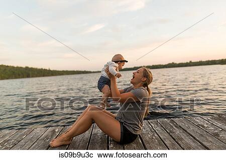 Stock Image - Mother sitting on lake pier holding up baby daughter. Fotosearch