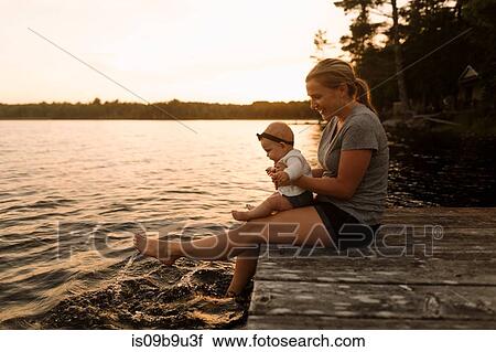 Stock Image - Mother sitting on pier with baby daughter, splashing feet in lake. Fotosearch