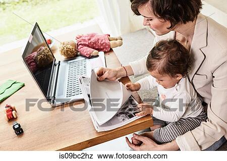 Stock Photo - Mother sitting with baby daughter, looking through paperwork, laptop on table in front of her. Fotosearch
