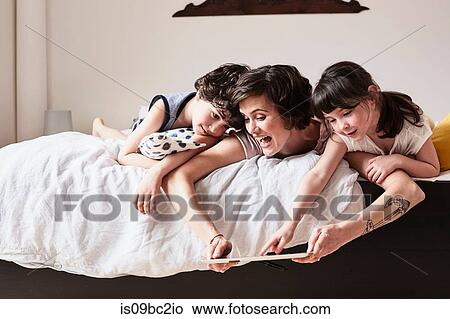 Picture - Mother, son and daughter lying on bed, using digital tablet. Fotosearch